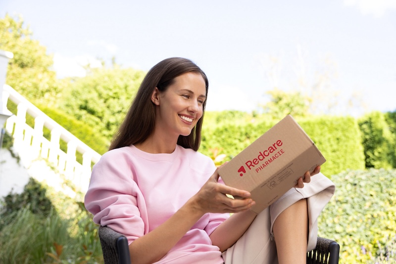 Une femme souriante est assise dans un jardin et tient un colis de Shop Apotheke.